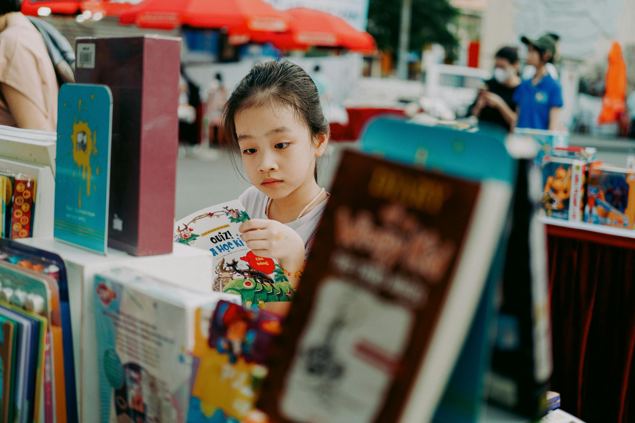 Curious young girl browsing books at an outdoor fair, surrounded by vibrant book covers.